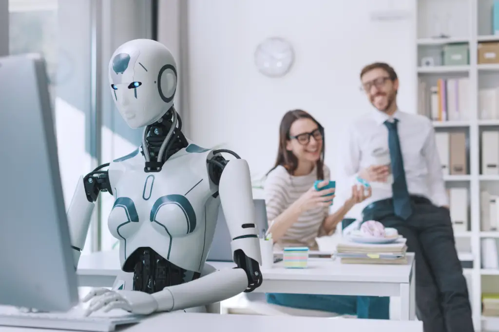 Robô com design moderno trabalhando em um computador dentro de um escritório, enquanto dois colegas humanos riem ao fundo. Representação da inteligência artificial no ambiente corporativo e a integração entre tecnologia e trabalho.