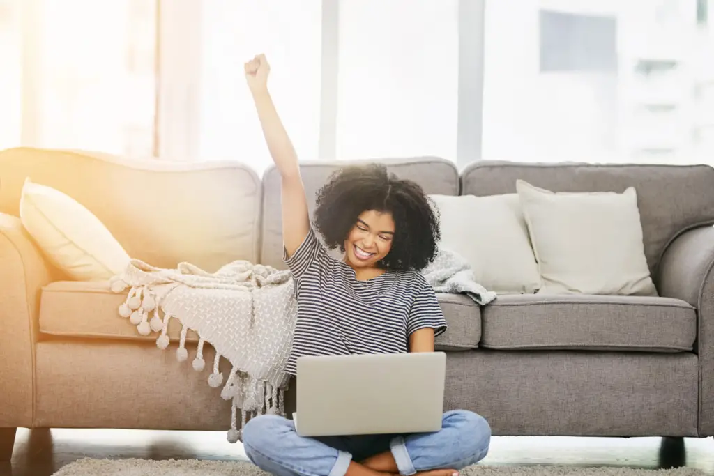 Mulher comemorando na frente do laptop Uma jovem feliz, sentada no chão da sala, levanta o braço em comemoração enquanto usa um laptop, sugerindo conquistas online, sucesso em negócios digitais ou boas notícias.